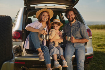 A family is gathered at an outdoor picnic, sitting on the back of their vehicle. They are sharing laughter, holding drinks, and enjoying each other's company against a beautiful backdrop.
