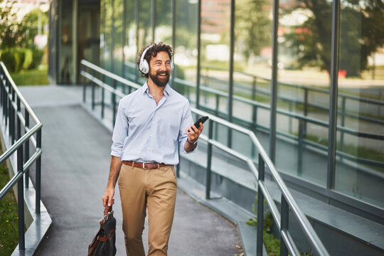 A young man dressed in business casual attire walks along a path outside a modern building, smiling while using his smartphone and wearing headphones. - Powered by Adobe