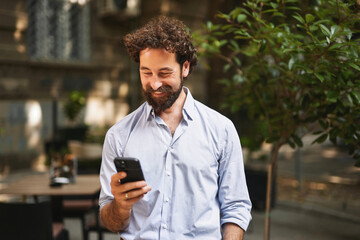 A man in a light blue shirt smiles at his smartphone, showcasing a positive interaction. He stands...