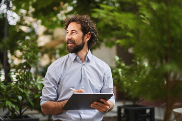 A man with a beard, dressed in a shirt, sits in an outdoor setting surrounded by lush greenery. He...