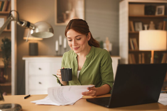 A woman dressed in a green shirt sits at a wooden desk, smiling as she sips from a black mug while reviewing paperwork in a comfortable home office filled with books.