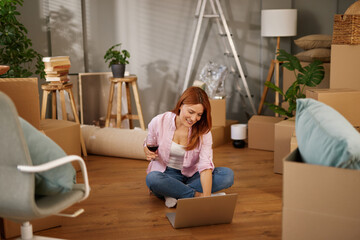 A woman sits on the floor surrounded by cardboard boxes, enjoying a drink and using her laptop as...