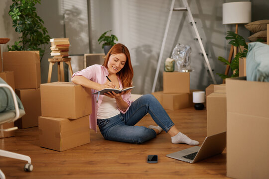 A woman sits on the floor surrounded by cardboard boxes, writing notes in a notebook. She is organizing her thoughts for the move while using a laptop close by. - Powered by Adobe