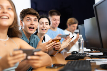 Positive guy student of computer courses sits among students and holds mobile phone in hands, plays game, tests new application