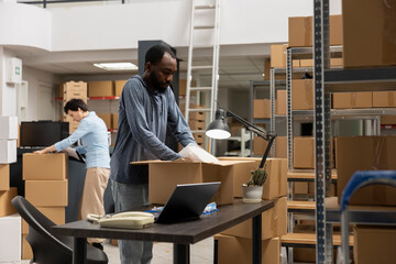 Black male executive manager uses laptop for order logistics in the storage room, working in a...