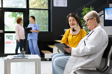 General practitioner assisting woman with cervical neck brace at hospital lobby, doing a routine...