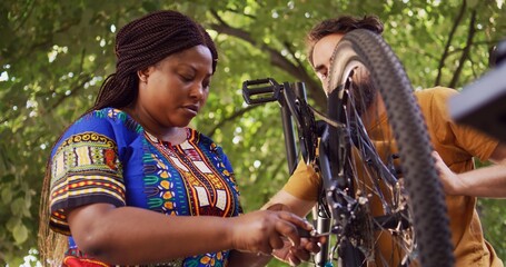 Active multiethnic couple servicing and repairing modern bicycle wheel outdoor. Caucasian boyfriend...