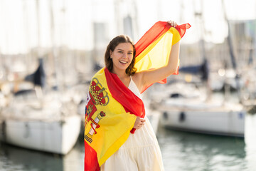 Young woman in summer dress wrapped in Spanish flag on sea port with yachts