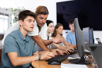 Obraz premium Interested young guy attending computer class, learning digital technology under guidance of positive adult male teacher standing nearby..