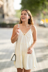 Cheerful girl in white dress walking along the street among architecture with a bag