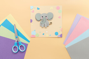 Gray elephant made from paper on square peach sheet surrounded by multicolored cut-out shapes, scissors, and pastel paper sheets on white background. flat lay. Animal craft project for kids. 