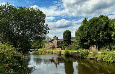 The Leeds and Liverpool Canal flows past an old stone house with leafy trees, mirrored in calm water beneath a dappled sky in Shipley, Yorkshire. UK