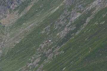 A red and white helicopter flies over a vast green mountain slope with a small cabin, conveying...
