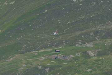A red and white helicopter flies over a vast green mountain slope with a small cabin, conveying...