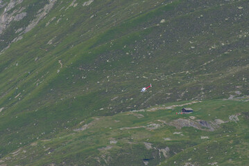 A red and white helicopter flies over a vast green mountain slope with a small cabin, conveying...