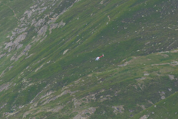 A red and white helicopter flies over a vast green mountain slope with a small cabin, conveying...