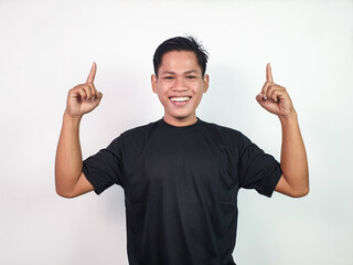 Smiling young Asian man in a black shirt, pointing up with both index fingers against a bright...