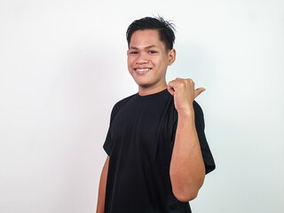 Happy young Asian man in a black shirt standing in a bright studio, smiling while pointing...