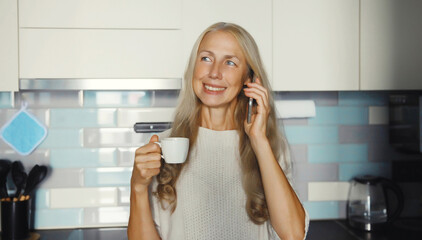 happy smiling woman talking on mobile phone enjoying hot tasty cup of coffee or tea in kitchen