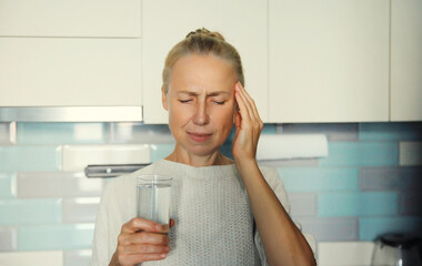 Sick upset mature woman suffering from headache holding glass of water, to take medicine at home