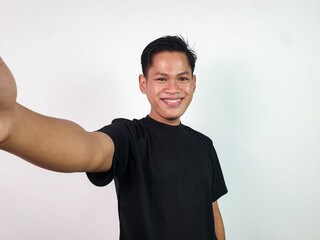 An enthusiastic young Asian man poses for a selfie with a cheerful expression, depicting joy and...