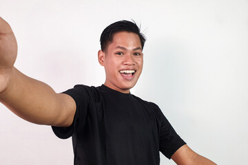 An enthusiastic young Asian man poses for a selfie with a cheerful expression, depicting joy and...