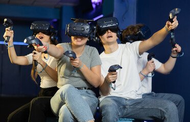Group of excited young people with handheld controllers in hands engaged in virtual world...