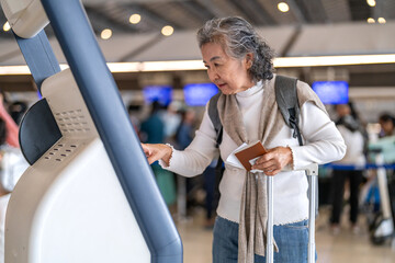 Asian senior woman traveler passenger with luggage using digital check in at airport kiosk, smart...