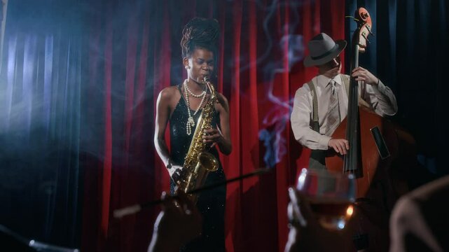 Front view shot of glamorous Black woman playing saxophone on stage while performing as bass duo in vintage jazz club with cigarette smoke in foreground