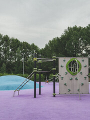 Colorful modern playground with climbing walls and structures on purple rubber surface and blue hill, surrounded by trees, concept of childhood activity and safe outdoor play