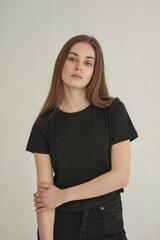 Obraz premium A studio portrait photograph against a plain off-white background. A young woman with long straight brown hair wearing a plain black crew neck t-shirt and dark jeans. 