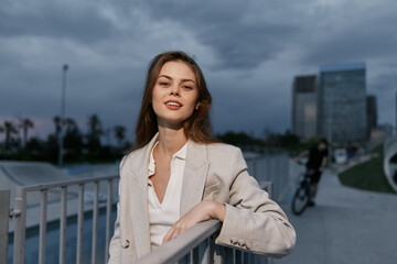 Young woman in beige blazer confidently posing outdoors with urban cityscape background cloudy sky...