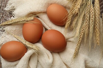 Raw eggs, fabric and spikes of wheat on wooden table, flat lay