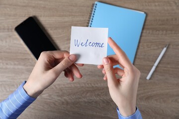 Woman holding paper note with word Welcome at wooden table, top view