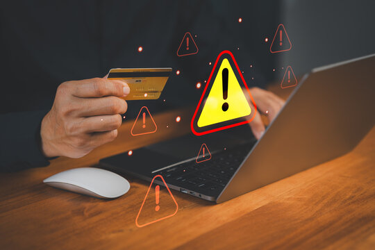 Customer hand holding a credit card while using a laptop with warning symbol, representing online payment risk, online shopping, financial fraud, or identity theft during digital transactions.