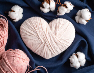 Heart-shaped pink yarn with cotton flowers and ball of yarn on soft blue textile background