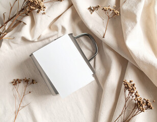 Metal mug and kraft envelope with blank tag on soft fabric surrounded by fresh green leaves