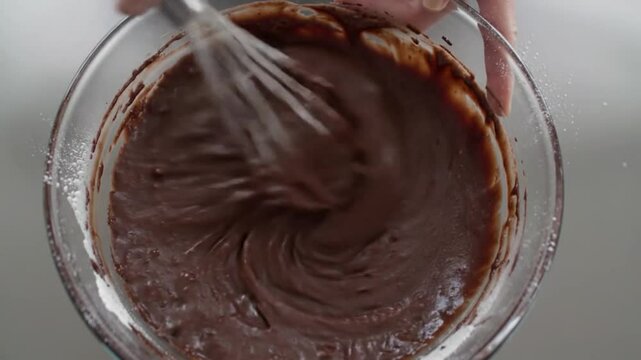 A large whisk mixing thick, smooth chocolate batter in a clean glass bowl, top-down view.