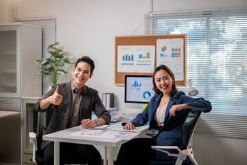 Two people in suits are sitting at a desk with a bulletin board behind them