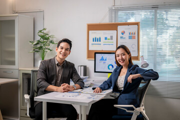 Two people in suits are sitting at a desk with a bulletin board behind them