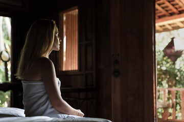 Peaceful morning moment. Woman in towel sits on bed looking outside. Calm thoughts, hotel stay, spa retreat, self-care, relaxation, wellness, natural light, wooden room, simple beauty.