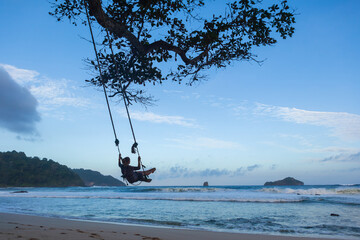 Happy girl have fun swinging high in mid air. Flying on rope swing on sea beach. Travel adventure on paradise tropical island. Family lifestyle, activity on summer vacation.