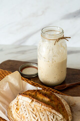 Crispy sourdough bread sits beside a glass jar filled with active starter, accompanied by a rustic...
