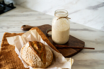 Crispy sourdough bread sits beside a glass jar filled with active starter, accompanied by a rustic...