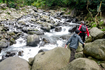 Obraz premium Two Hikers Navigate A Rocky River Stepping Carefully On Large Boulders Amidst Lush Greenery