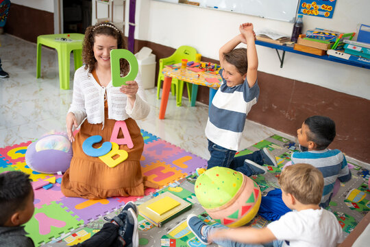 Preschool teacher holds colorful foam letters while sitting with a diverse group of children on the classroom floor. Early childhood education, alphabet learning, fun interactive play in kindergarten