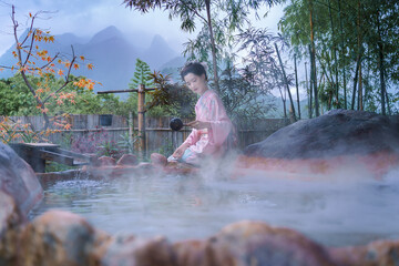 A beautiful Asian woman in a Traditional Furisode Kimono sits and relaxes by an outdoor Onsen Hot Spring. Japanese traditional cultural costume.