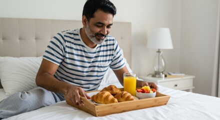 Man in bed with breakfast tray containing croissants fruit and orange juice in a bright bedroom setting