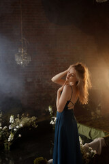 Young Caucasian woman in blue dress posing with raised arms, looking away. Front view. Vintage room with brick wall, crystal chandelier, flowers. Concepts: elegance, beauty, nostalgia, romance.
