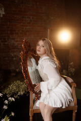 Young Caucasian woman with long blonde hair, in white dress, rear view, sits by a vintage mirror in a dark moody room with brick wall and flowers. Concepts: reflection, solitude, fantasy, dream.